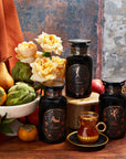 A rustic setup with three black spice jars labeled "Serpise," "Virtus," and "Libra," surrounded by a bowl of fruits including pears, pomegranates, and artichokes. Orange roses and a glass of tea in a clear teacup with a saucer complete the scene.