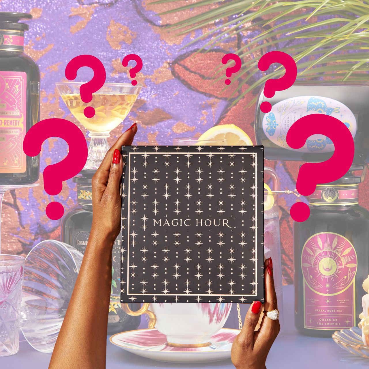 Two hands present a box labeled with "Black Friday Mystery Box," adorned with a star design, suggesting the excitement of its contents. The backdrop showcases vibrant bottles, elegant glassware, and palm leaves among prominent red question marks, alluding to the premium teas and tea accessories within.