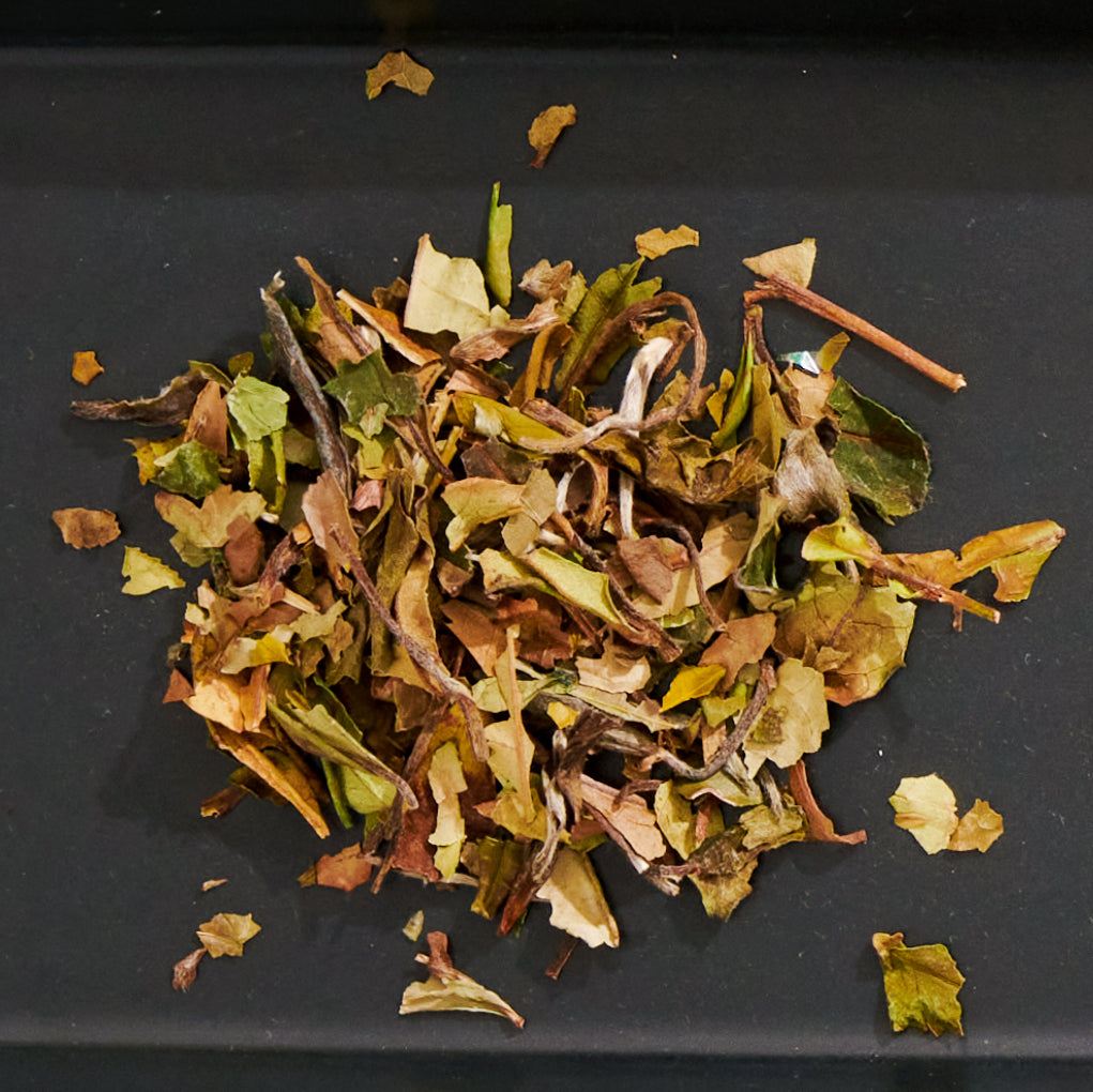 A close-up image of loose-leaf Bai Mudan White Tea by Magic Hour. The handcrafted blend includes various dried leaves and stems in shades of green, brown, and yellow, spread across a dark surface. The white tea leaves appear to be a mix of different sizes and shapes.