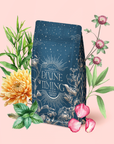 A blue floral coffee bag labeled "Divine Timing Balance" sits among green leaves, a yellow chrysanthemum, pink petals, mint herbs, and purple thistles—creating a balanced blend for women on a soft pink background.