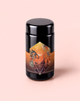 A black cylindrical container adorned with an orange, geometric label featuring abstract illustrations and text sits against a pale pink background. The design evokes the lively character of Bali Bliss: Coconut-Lime & Ginger-Melon White Tea, a blend inspired by island flavors—crafted from select white teas highlighted by bright tropical notes.
