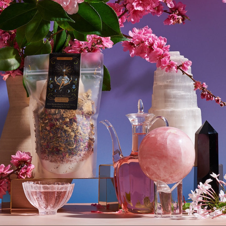 A stunning arrangement featuring the "High Priestess Bath Tea" from Magic Hour, pink cherry blossoms in a vase, a glass pitcher with pink liquid, crystal balls, and a black crystal obelisk, all set against a gradient purple and blue background that channels divine feminine energy.