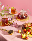 The Breakfast with Benefits Bundle features a glass teapot of red tea, two teacups, fresh fruit plate, and a small dish of loose tea leaves, all beautifully accented with purple flowers against a pink backdrop.