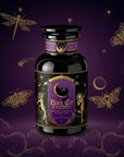 A dark glass jar labeled “Black Cat Brownie: Decadent Chocolate Puerh Blend with Vanilla & Hazelnut” sits on a purple background decorated with gold illustrations of moths, a dragonfly, stars, and clouds. This luxurious blend combines rich chocolate notes with smooth organic Pu-erh tea, complemented by hints of vanilla and hazelnut for an indulgent treat.