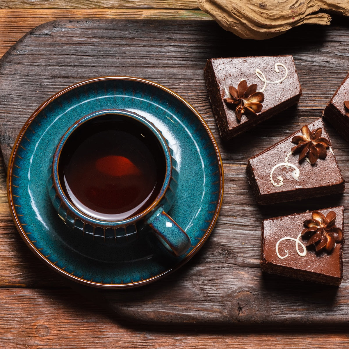 A teal cup of chocolate black tea on a matching saucer sits on a wooden table beside three Black Cat Brownies, each topped with flower-shaped frosting and gold accents.