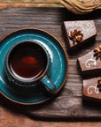 A teal cup of chocolate black tea on a matching saucer sits on a wooden table beside three Black Cat Brownies, each topped with flower-shaped frosting and gold accents.