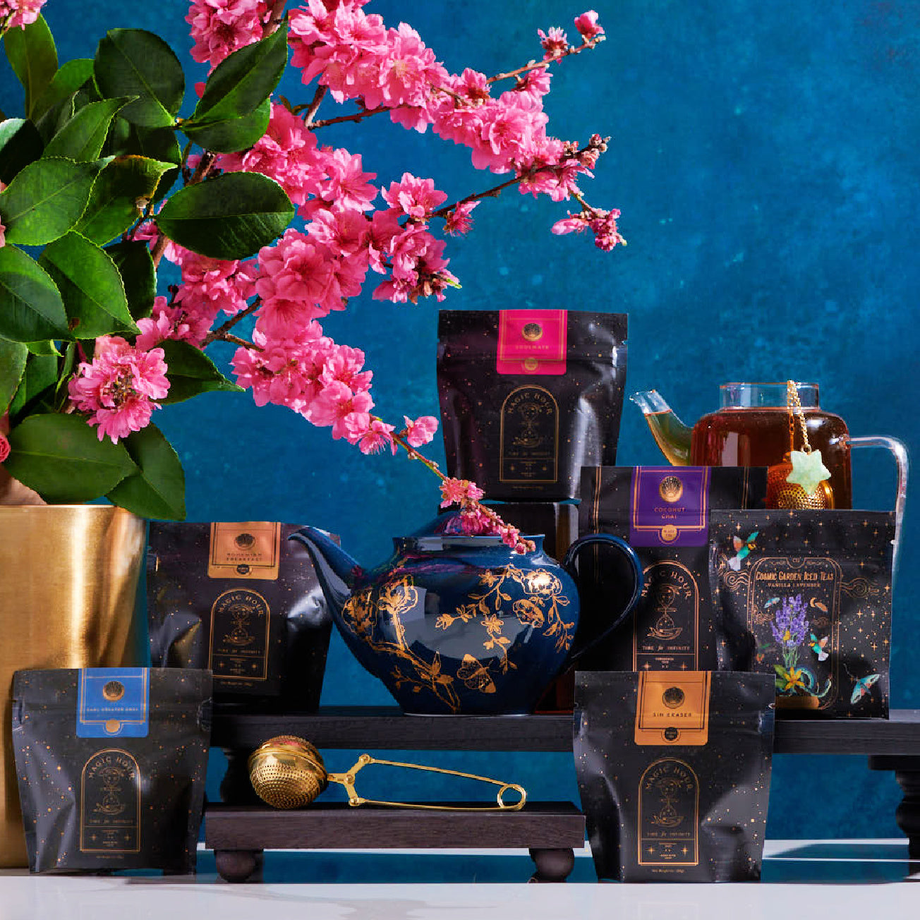 A colorful display features various bags of Magic Hour's Black Tea Magic Sampler Box - Bestselling Organic Black Teas for Energy & Vitality on a dark wooden rack, with a dark blue teapot adorned with golden patterns in the center. A gold tea infuser and a glass teapot with organic tea are nearby. Pink cherry blossoms in a pot enhance the vibrant setting.