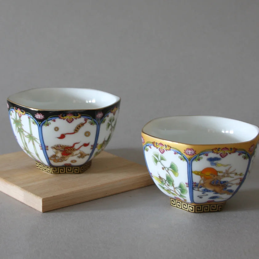 The Hexagon Cups from Eastern Elm, displayed on a wooden surface, beautifully capture the elegance of traditional Chinese folk art. The cup on the left showcases an intricate black, white, and gold design adorned with floral and mythical elements, while the cup on the right features high-quality porcelain with exquisite white, blue, and gold bird motifs. Both cups are elegantly finished with gold trims.