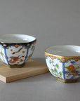 Displayed on a light wooden block are the Hexagon Cups - Set of 2 by Eastern Elm, two ornate porcelain teacups featuring intricate floral and nature designs. One cup showcases a predominantly black and white motif, while the other boasts a white base with gold accents. These high-quality porcelain cups have traditional Asian-style patterns, making them perfect for tea ceremonies.