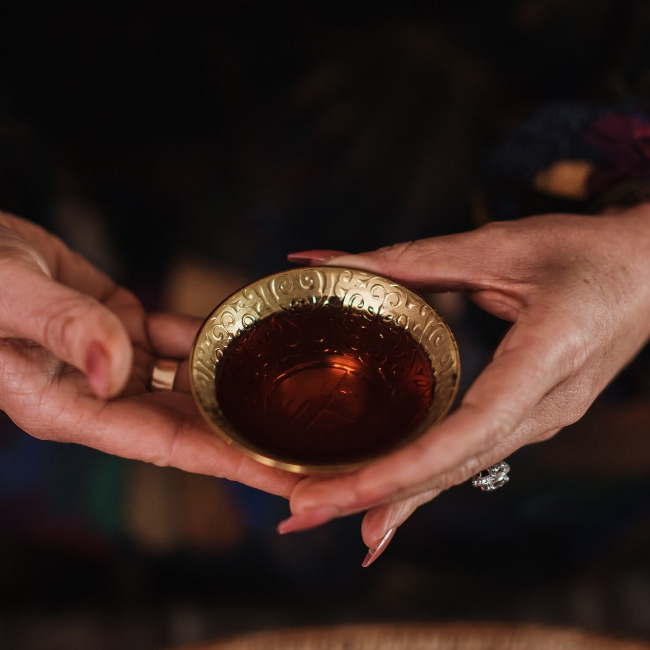 Close-up of a person's hands holding a small, ornately decorated brass cup filled with tea. The person's fingers have polished nails, and they are wearing a beaded bracelet and a ring with a large stone. The background is softly blurred and dark.