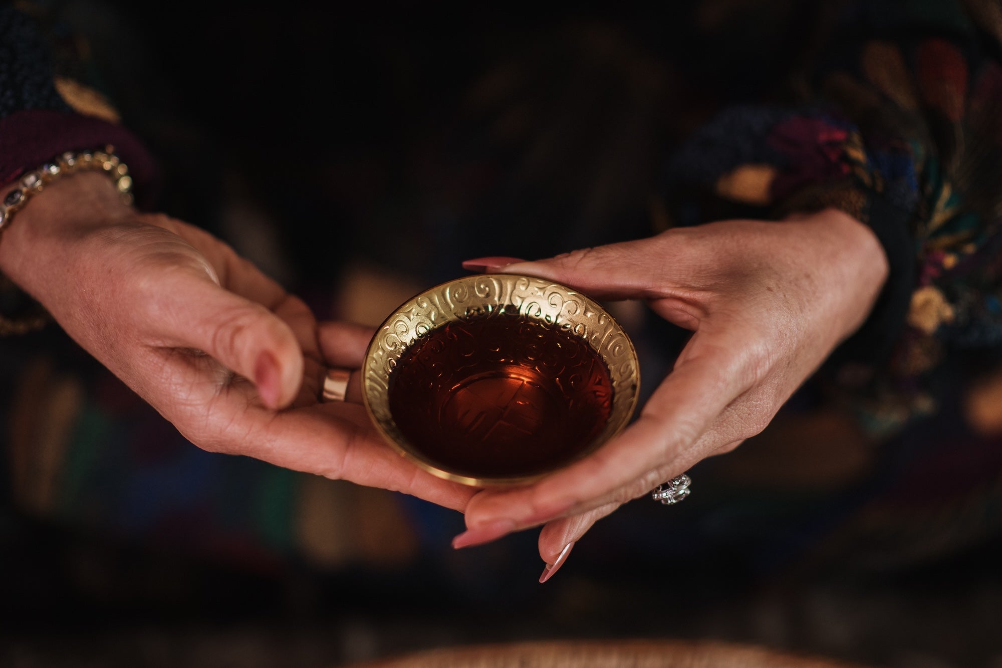 Close-up of a person's hands holding a small, ornately decorated brass cup filled with tea. The person's fingers have polished nails, and they are wearing a beaded bracelet and a ring with a large stone. The background is softly blurred and dark.