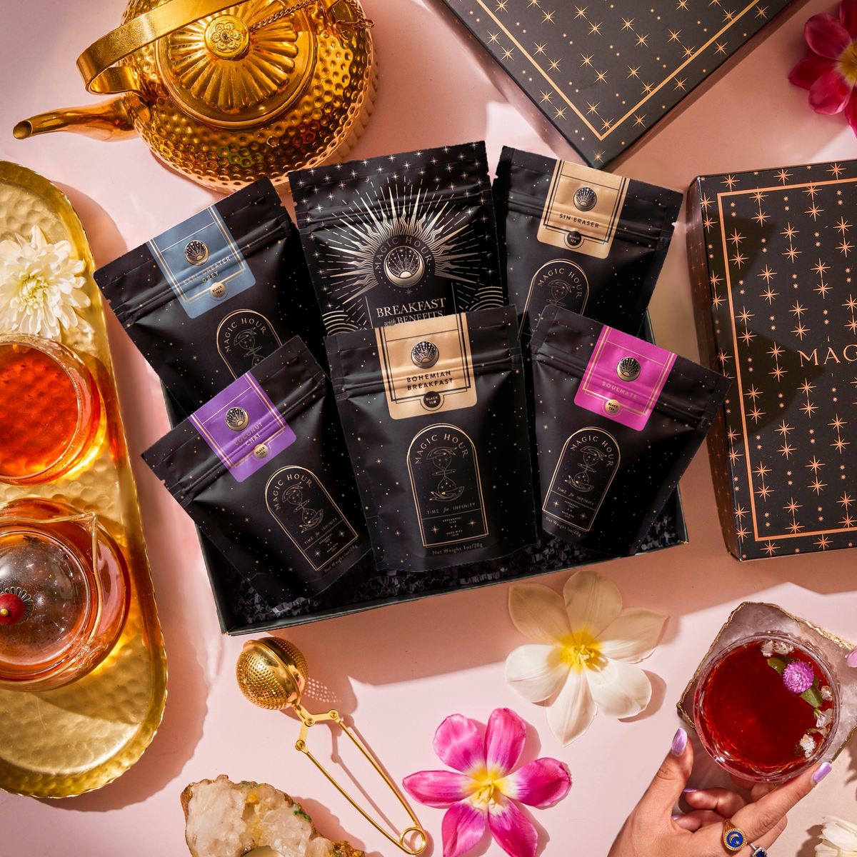 A Black Tea Sampler Box: Bestselling Organic Black Teas for Energy & Vitality containing six tea pouches, surrounded by a gold teapot, glass teacups, a tea infuser, and fresh flowers.