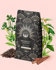 A decorative bag labeled "Blood Sugar Balance: Breakfast with Benefits" stands surrounded by aromatic herbs, Assam black tea leaves, and fragrant cinnamon sticks on a light pink background.