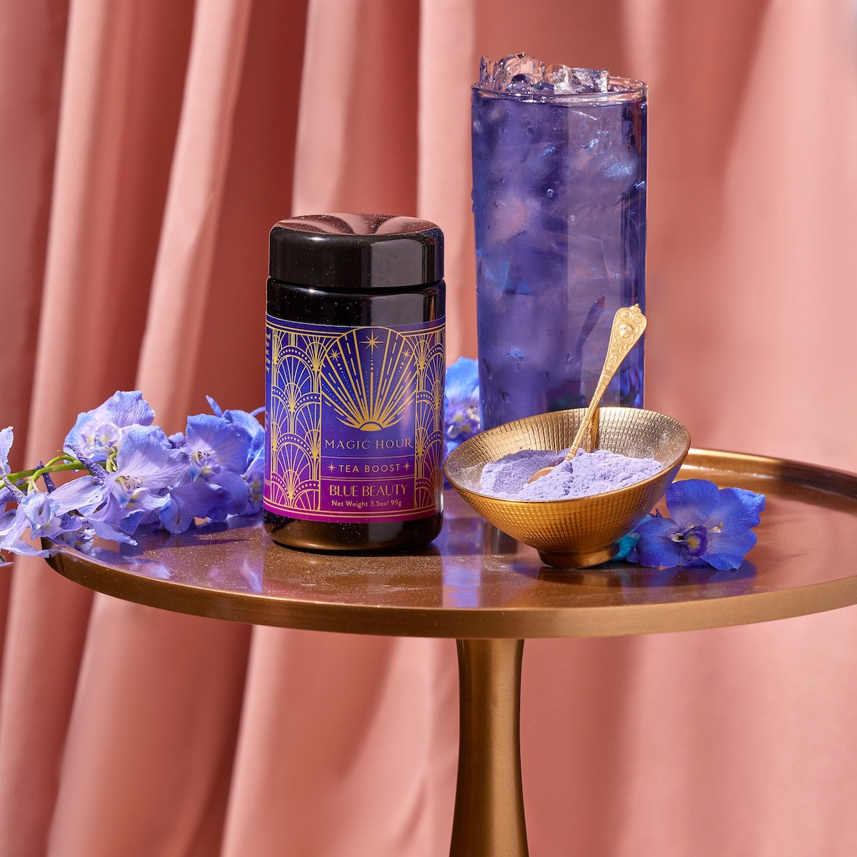 A container of Blue Beauty Tea Boost is placed on a round gold table along with a scoop of purple tea powder in a bowl with a spoon. A tall glass of iced blue tea and blue flowers are also on the table, set against soft pinkish drapes in the background.