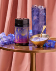 A container of Blue Beauty Tea Boost is placed on a round gold table along with a scoop of purple tea powder in a bowl with a spoon. A tall glass of iced blue tea and blue flowers are also on the table, set against soft pinkish drapes in the background.