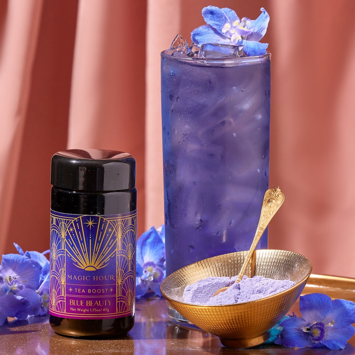 A tall glass filled with blue iced tea is garnished with purple flowers. Beside it is a jar labeled &quot;Blue Beauty Tea Boost&quot; and a gold bowl containing purple powder with a gold spoon. The background features a pink curtain.