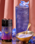 A tall glass filled with blue iced tea is garnished with purple flowers. Beside it is a jar labeled "Blue Beauty Tea Boost" and a gold bowl containing purple powder with a gold spoon. The background features a pink curtain.