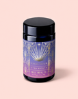 A black canister of Magic Hour’s Blue Beauty: Butterfly Pea Flower Powder Boost tea with an art deco label in blue, gold, and purple—adorned with sun and starburst motifs—set against a soft pink background. This blend features butterfly pea flower powder, celebrated for supporting collagen production and promoting radiant skin.