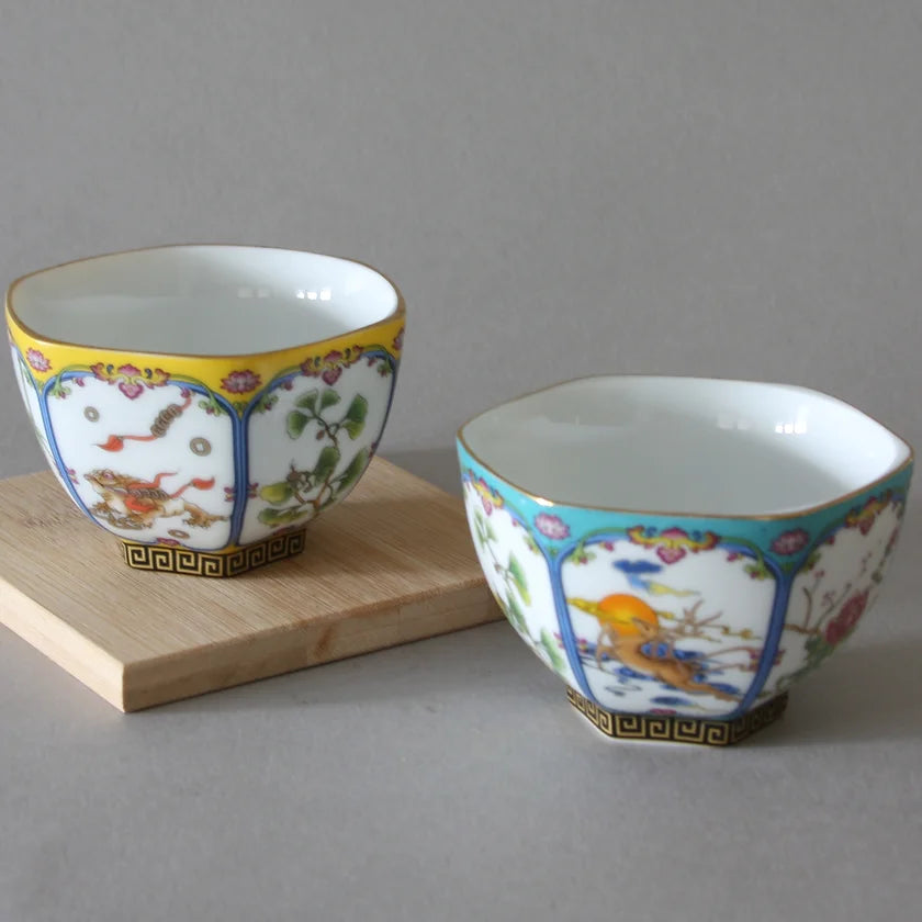 The Hexagon Cups - Set of 2 from Eastern Elm, intricately decorated with one cup sporting a yellow upper rim and the other a blue upper rim, sit on a light wooden base against a gray background. Reminiscent of traditional Chinese folk art, each cup features floral and animal motifs adorned with gold accents.