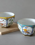 The Hexagon Cups - Set of 2 from Eastern Elm, intricately decorated with one cup sporting a yellow upper rim and the other a blue upper rim, sit on a light wooden base against a gray background. Reminiscent of traditional Chinese folk art, each cup features floral and animal motifs adorned with gold accents.