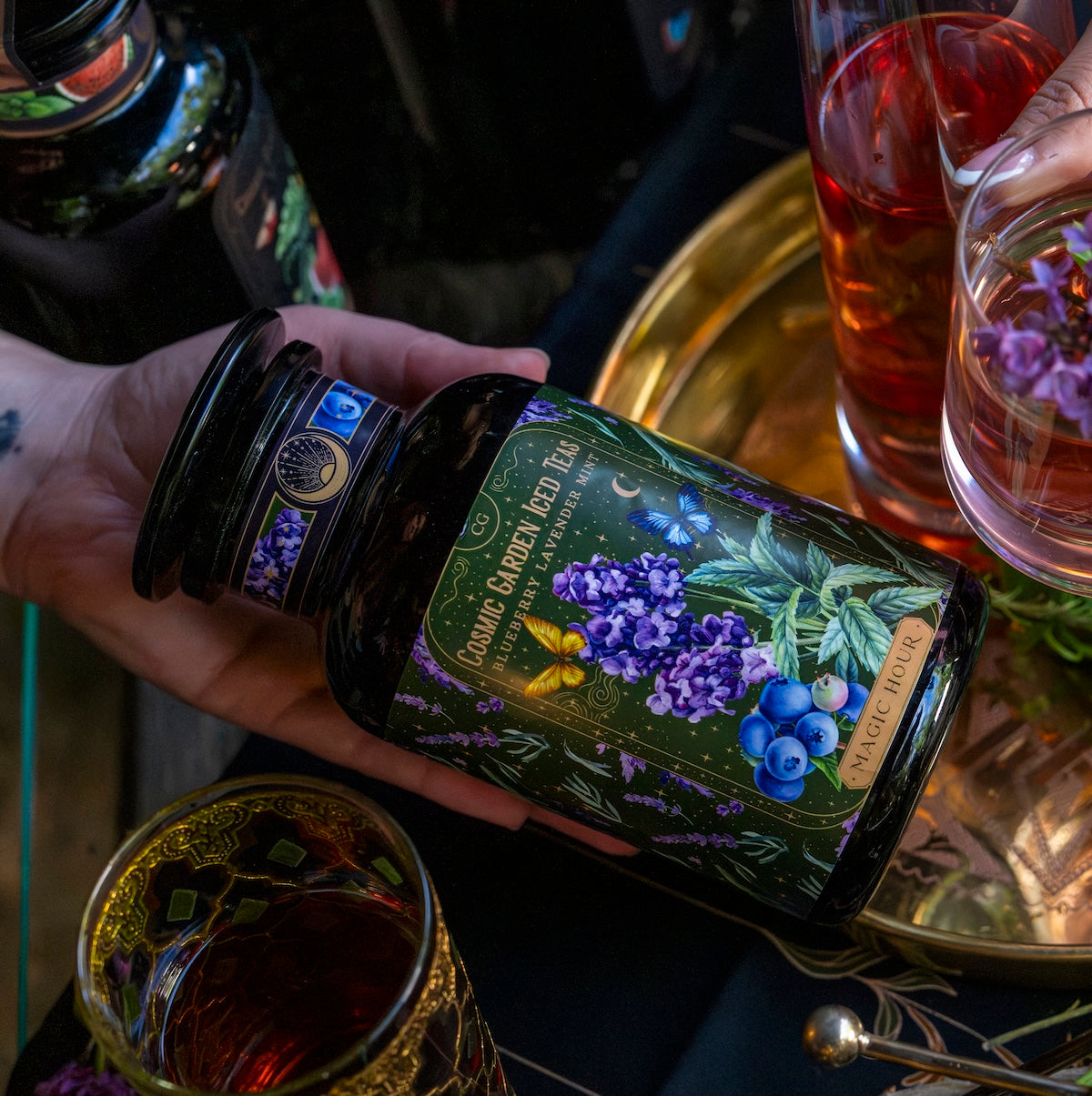 A hand holds a jar labeled &quot;Magic Hour&#39;s Blueberry Lavender Mint: Cosmic Garden Iced Tea&quot; with an intricate design featuring blueberries and purple flowers. The jar, brimming with fragrant loose leaf tea, is held over a gold tray surrounded by ornate glasses of beverages. The scene is dimly lit, creating a cozy and enchanting atmosphere.