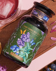 A jar of Blueberry Lavender Mint: Cosmic Garden Iced Tea sits on a wooden tray. The jar features floral and berry designs. Surrounding the jar are petals, loose leaf tea mix, blueberries, and a glass of iced tea with a pinkish hue. This Magic Hour product promises an enchanting sip.