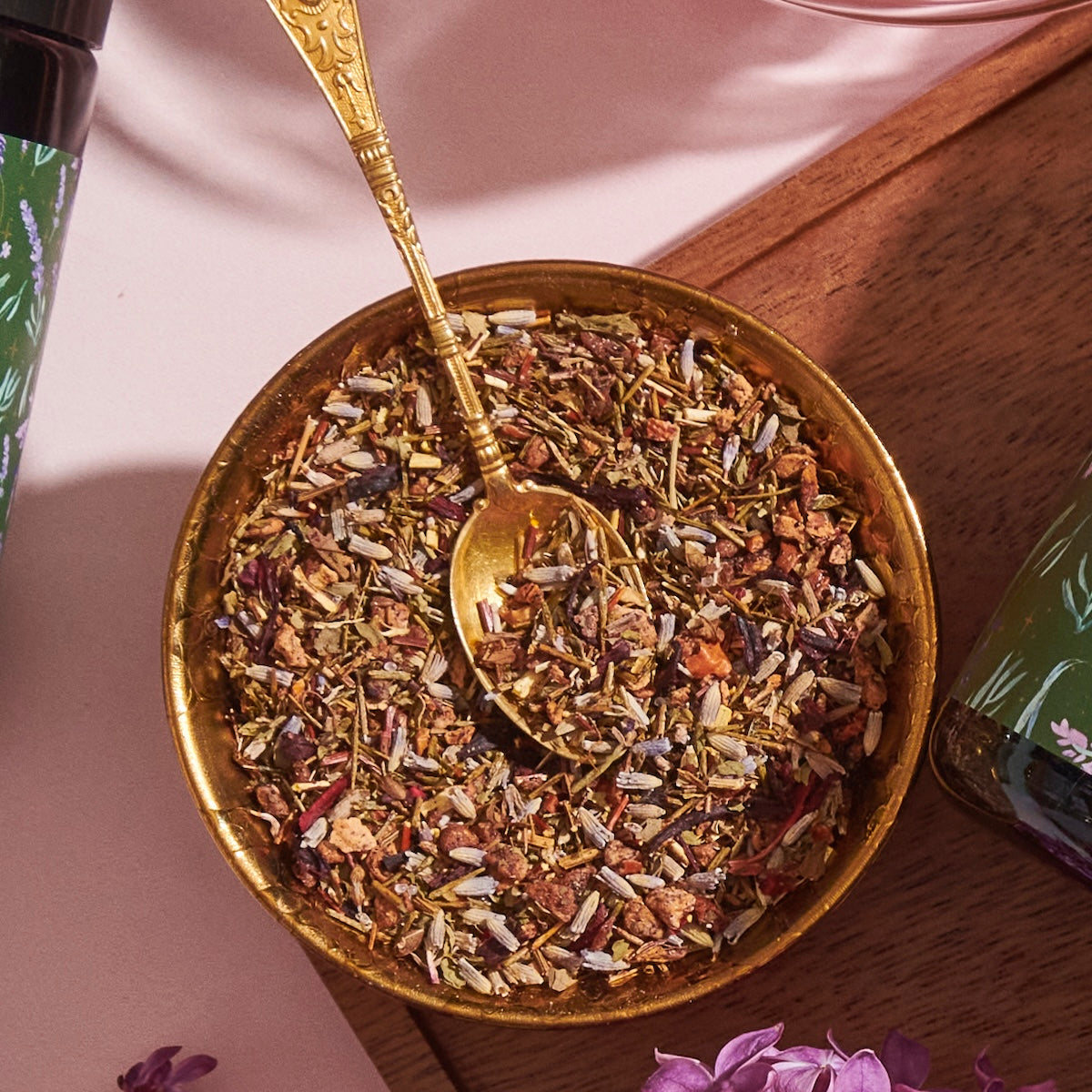 A brass bowl filled with a colorful mixture of dried herbs and spices, reminiscent of loose leaf tea, is accompanied by an ornate brass spoon. The bowl is set on a wooden surface with a bottle of Magic Hour&#39;s Blueberry Lavender Mint: Cosmic Garden Iced Tea featuring floral designs partially visible to the left.