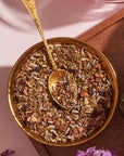 A brass bowl filled with a colorful mixture of dried herbs and spices, reminiscent of loose leaf tea, is accompanied by an ornate brass spoon. The bowl is set on a wooden surface with a bottle of Magic Hour's Blueberry Lavender Mint: Cosmic Garden Iced Tea featuring floral designs partially visible to the left.