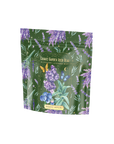 A green metallic tea packaging with colorful illustrations of lavender, mint leaves, a butterfly, and blueberries. It reads "Blueberry Lavender Mint: Cosmic Garden Iced Tea." The brand "Magic Hour" is prominently displayed at the bottom of the pouch.