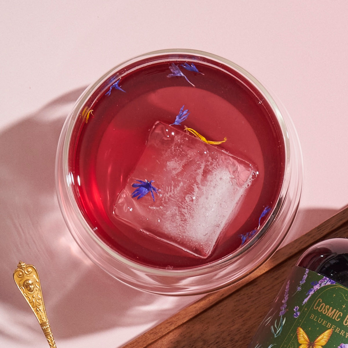 A bright pink beverage in a clear glass is garnished with a large, clear ice cube and small blue and yellow flower petals. The drink, crafted from Blueberry Lavender Mint: Cosmic Garden Iced Tea by Magic Hour, is placed on a light pink surface near a golden spoon and a partially visible bottle with a colorful label.