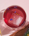 A bright pink beverage in a clear glass is garnished with a large, clear ice cube and small blue and yellow flower petals. The drink, crafted from Blueberry Lavender Mint: Cosmic Garden Iced Tea by Magic Hour, is placed on a light pink surface near a golden spoon and a partially visible bottle with a colorful label.