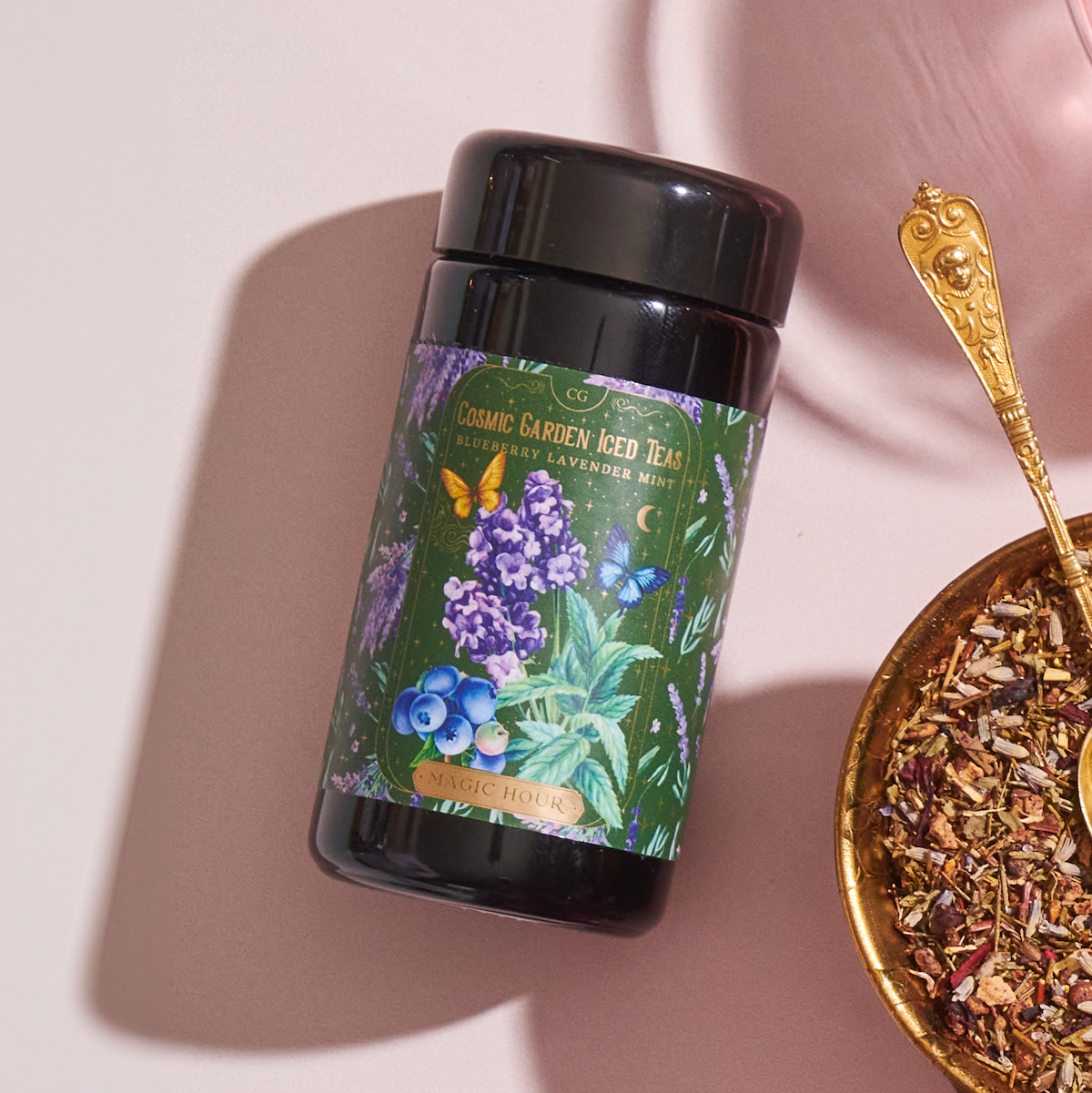 An ornate gold spoon is filled with loose leaf tea, positioned next to a container of Magic Hour’s Blueberry Lavender Mint: Cosmic Garden Iced Tea. The container has a colorful label featuring illustrations of butterflies and flowers. The scene is set against a soft pink background, exuding an enticing allure.