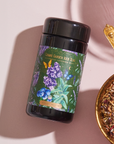 An ornate gold spoon is filled with loose leaf tea, positioned next to a container of Magic Hour’s Blueberry Lavender Mint: Cosmic Garden Iced Tea. The container has a colorful label featuring illustrations of butterflies and flowers. The scene is set against a soft pink background, exuding an enticing allure.