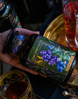 A hand holds a jar labeled "Magic Hour's Blueberry Lavender Mint: Cosmic Garden Iced Tea" with an intricate design featuring blueberries and purple flowers. The jar, brimming with fragrant loose leaf tea, is held over a gold tray surrounded by ornate glasses of beverages. The scene is dimly lit, creating a cozy and enchanting atmosphere.