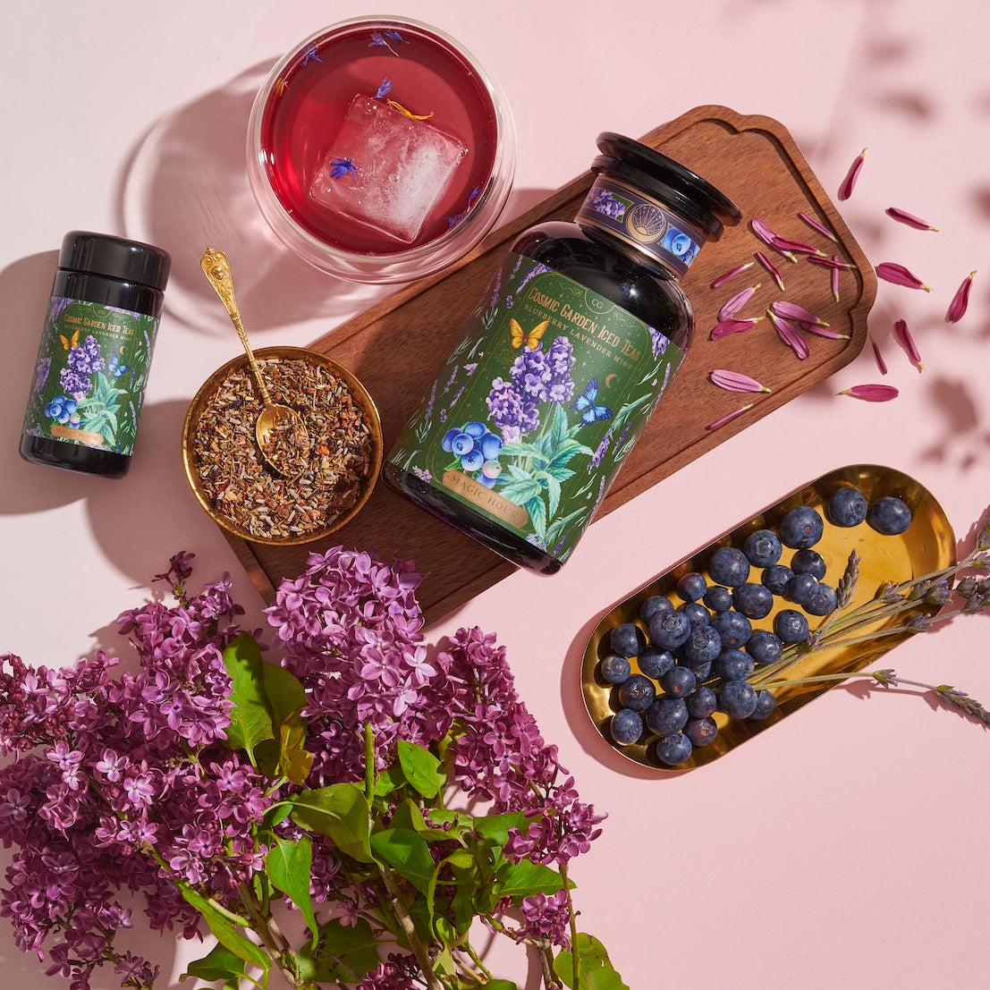 A vibrant arrangement featuring a large jar, a small bottle, a cup of Blueberry Lavender Mint: Cosmic Garden Iced Tea by Magic Hour with an ice cube, a bowl of loose leaf tea with a gold spoon, a bunch of purple flowers, and a gold tray of blueberries during the magic hour. The jars have botanical labels, and petals are scattered on a wooden board.