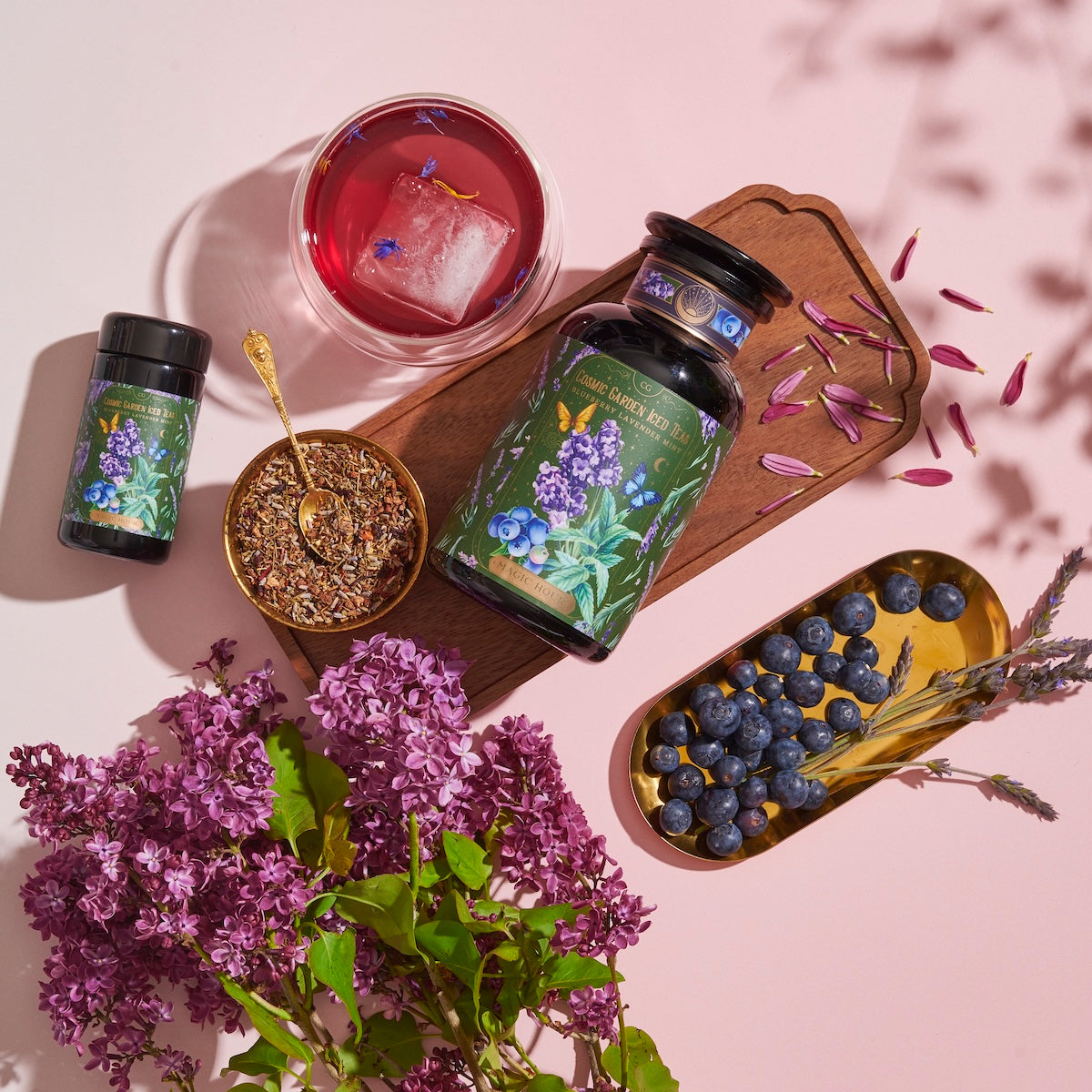 A vibrant arrangement featuring a large jar, a small bottle, a cup of Blueberry Lavender Mint: Cosmic Garden Iced Tea by Magic Hour with an ice cube, a bowl of loose leaf tea with a gold spoon, a bunch of purple flowers, and a gold tray of blueberries during the magic hour. The jars have botanical labels, and petals are scattered on a wooden board.