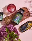 A vibrant arrangement featuring a large jar, a small bottle, a cup of Blueberry Lavender Mint: Cosmic Garden Iced Tea by Magic Hour with an ice cube, a bowl of loose leaf tea with a gold spoon, a bunch of purple flowers, and a gold tray of blueberries during the magic hour. The jars have botanical labels, and petals are scattered on a wooden board.