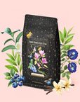 A tea bag labeled “Blueberry Cheesecake Oolong Iced Tea,” adorned with a starry design and floral illustrations, rests among blueberries, leaves, magnolia flowers, and morning glories on a pastel pink background—evoking an enchanting cosmic garden scene.