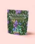 A green resealable pouch features lavender, butterflies, blueberries, and gold accents. The label says "Blueberry Lavender Mint: Cosmic Garden Iced Tea" on pink. This sugar-free blend is antioxidant-rich and delicious.