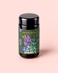 A canister of Blueberry Lavender Mint: Cosmic Garden Iced Tea from Magic Hour, decorated with a vibrant label displaying lavender, mint, and blueberries against a green background, rests on a pale pink surface. This sugar-free blend is ideal for savoring an antioxidant-rich iced tea at any time of day.