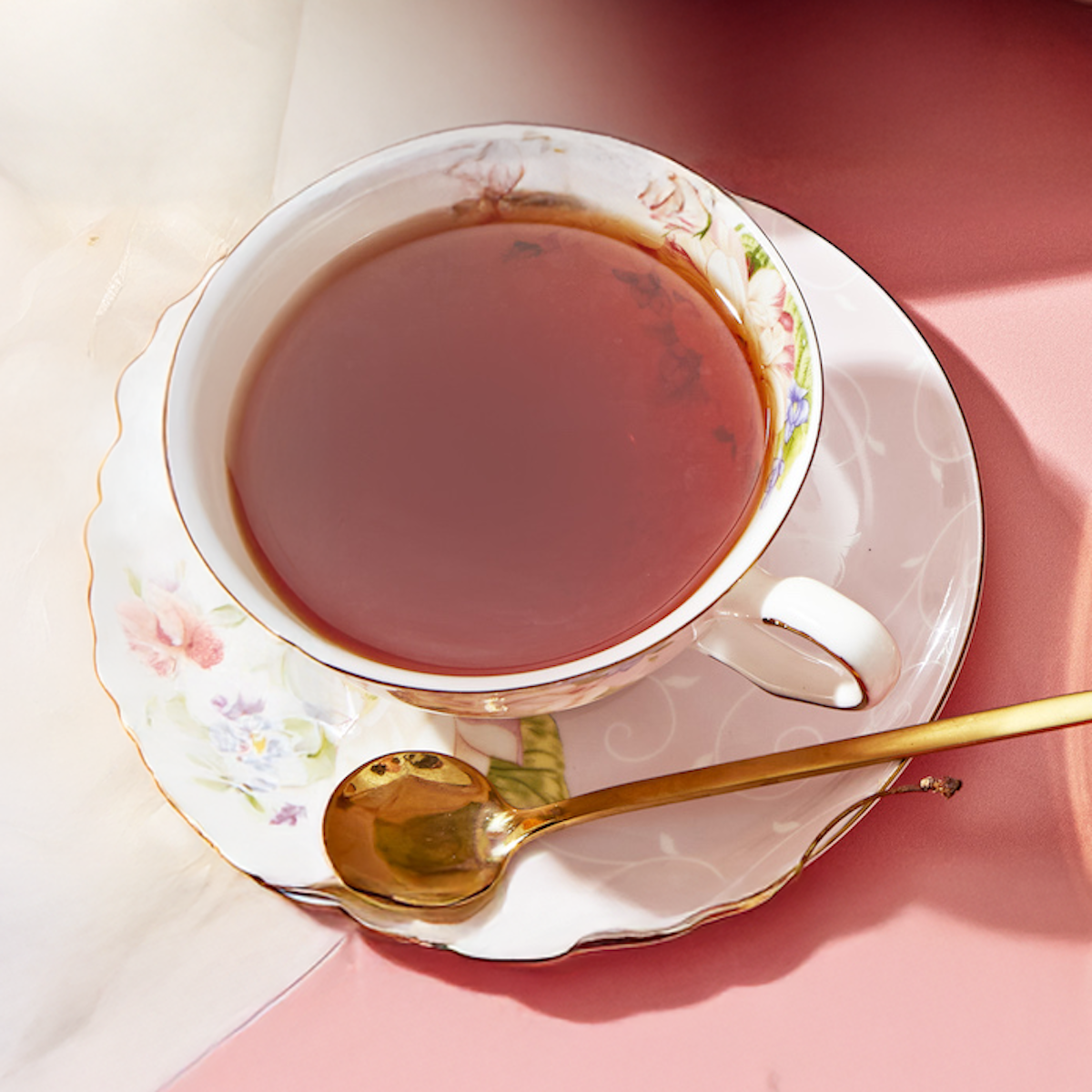 A Bohemian Breakfast Black Tea Bag steeps in a floral teacup on a matching saucer with a gold spoon, set atop a pink surface and soft white cloth—a stylish, organic coffee alternative.