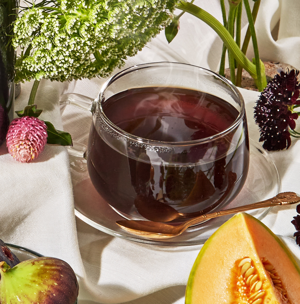 A glass cup of Bohemian Breakfast Black Tea sits on a white cloth with a saucer and gold spoon, surrounded by fresh flowers, fig, and cantaloupe. Steam rises from the tea, offering cozy comfort as a perfect coffee alternative.
