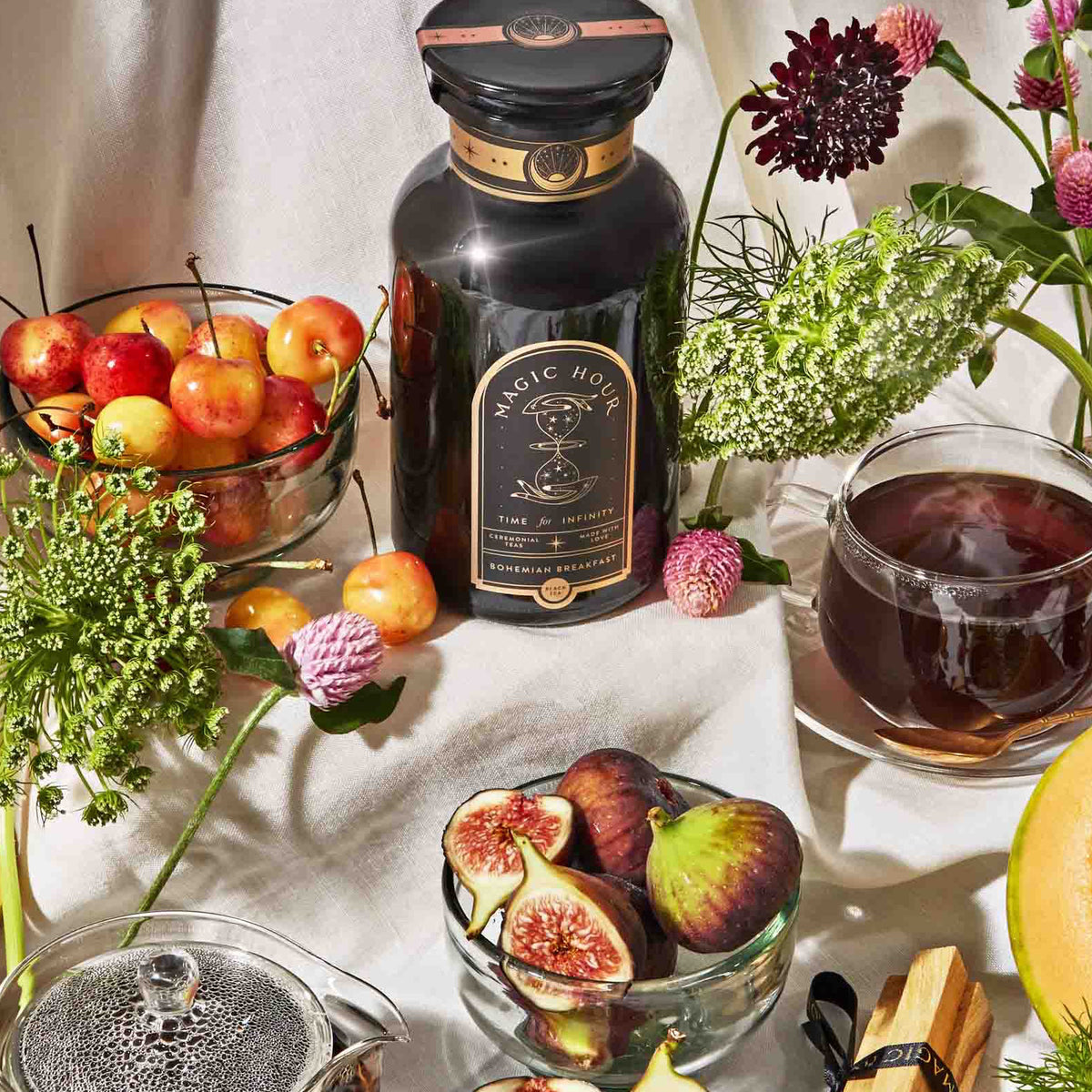 A dark apothecary jar of Best Sellers of Magic Hour Trio Box Bohemian Breakfast tea surrounded by a bowl of cherries, a bowl of sliced figs, a cup of tea, and pieces of cantaloupe.