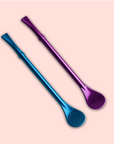 Two Aurora Bombilla stainless steel tea straws—one blue, one purple, each with a flat perforated end—lie diagonally on a light pink background, offering a stylish and sustainable way to sip your favorite drinks.