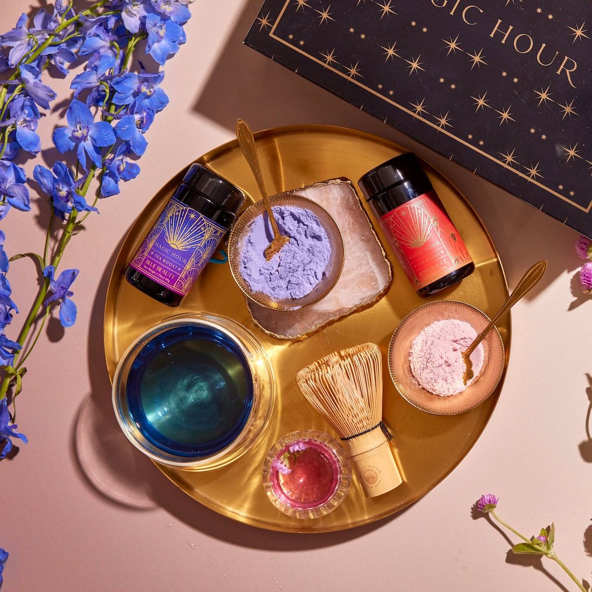 The Hydrate & Glow Tea Boost Bundle features a golden tray showcasing two small glass jars with vibrant blue and red labels, accompanied by a whisk, small containers filled with purple and pink powders, and a glass labeled "Blue Beauty Boost" containing a blue liquid. A tiny pink flower adds an elegant touch, while a decorative box and purple flowers are artfully arranged beside the tray.