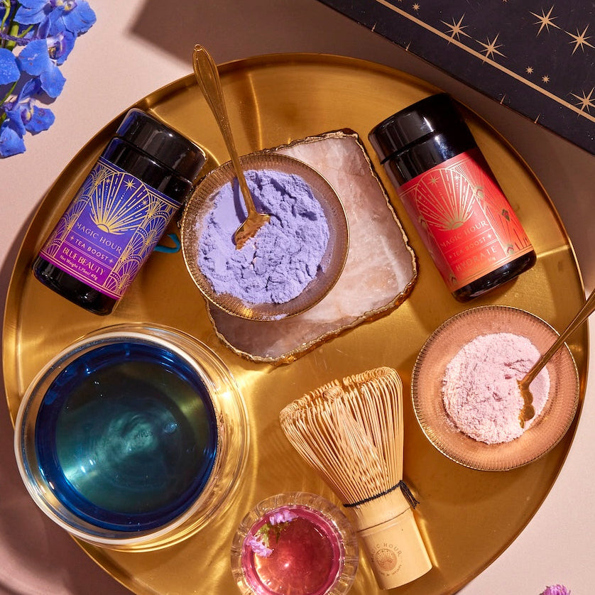 The Hydrate & Glow Tea Boost Bundle features a golden tray showcasing two small glass jars with vibrant blue and red labels, accompanied by a whisk, small containers filled with purple and pink powders, and a glass labeled "Blue Beauty Boost" containing a blue liquid. A tiny pink flower adds an elegant touch, while a decorative box and purple flowers are artfully arranged beside the tray.
