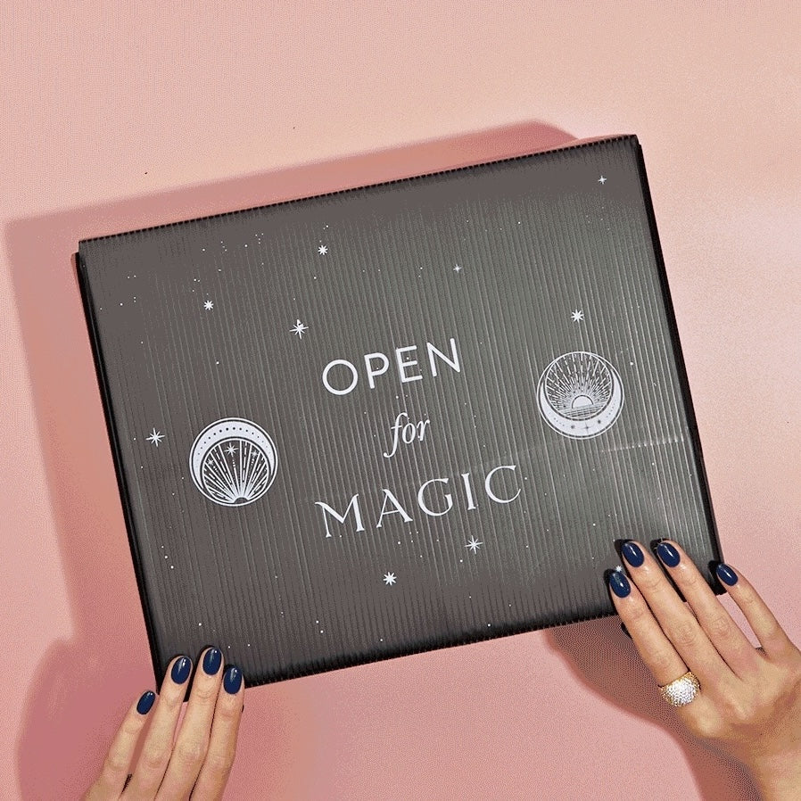 A person with dark blue nail polish holds a black box labeled "Open for Magic," which rests on a pink surface. The box, adorned with seashell and starry circle symbols, hints at enchanting experiences within. It may be the School of Magic Tea Subscription Box - Petite Apothecary Jar.