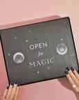 A person with dark blue nail polish holds a black box labeled "Open for Magic," which rests on a pink surface. The box, adorned with seashell and starry circle symbols, hints at enchanting experiences within. It may be the School of Magic Tea Subscription Box - Petite Apothecary Jar.