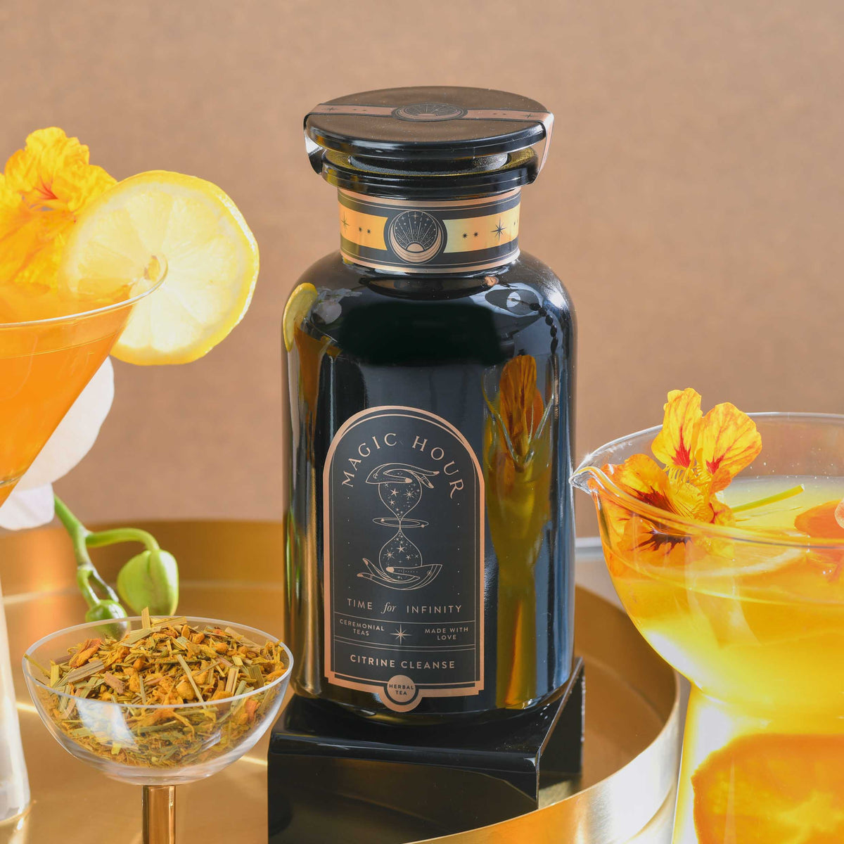 A black bottle labeled “Citrine Cleanse™ Herbal Tea” sits on a gold tray, surrounded by caffeine-free herbal tea, yellow flowers, a lemon slice, and two colorful drinks. The warm beige background highlights this detoxifying elixir infused with organic Hawaiian Ginger.