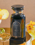 A black bottle labeled “Citrine Cleanse™ Herbal Tea” sits on a gold tray, surrounded by caffeine-free herbal tea, yellow flowers, a lemon slice, and two colorful drinks. The warm beige background highlights this detoxifying elixir infused with organic Hawaiian Ginger.